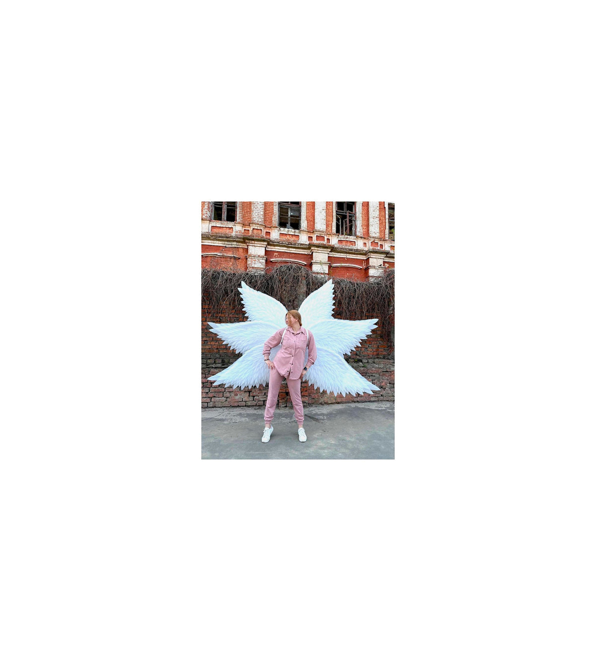 May include: A person wearing a pink outfit with white angel wings standing in front of a brick building.
