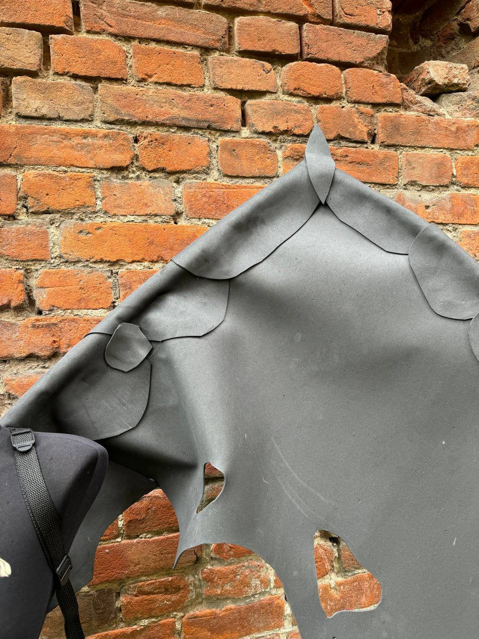 May include: A black fabric with a unique design of cutouts in the shape of leaves. The fabric is hanging against a brick wall.