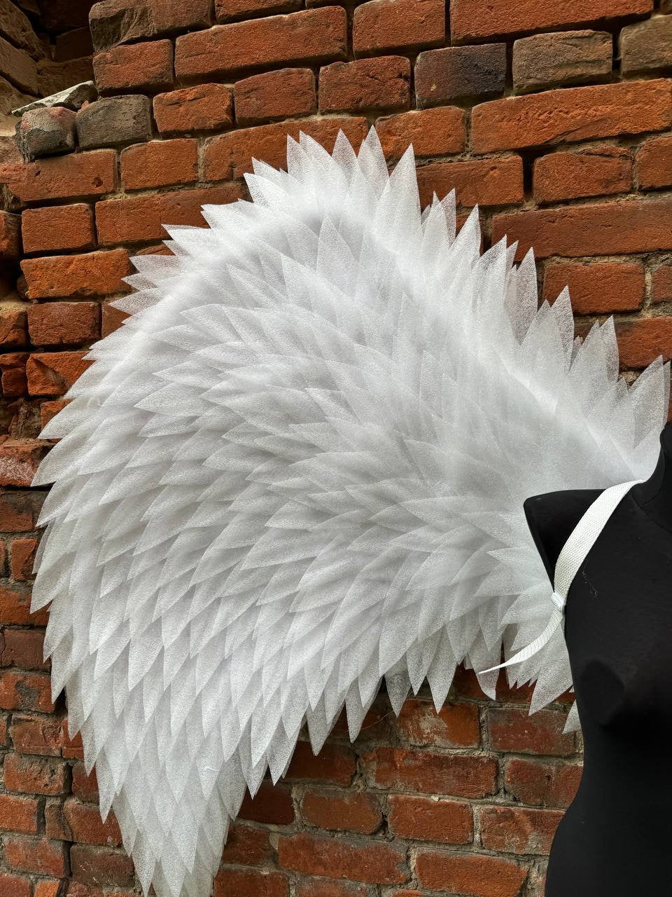 May include: A pair of large, white angel wings made of layered fabric. The wings are attached to a white strap and are leaning against a brick wall.