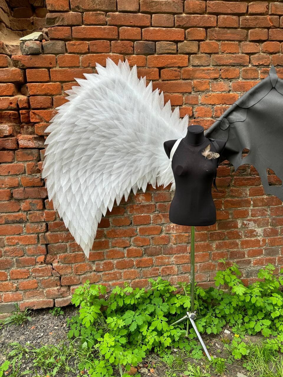 May include: A white angel wing and a black devil wing are attached to a black mannequin torso. The mannequin is on a metal stand with three legs. The wings are against a brick wall.