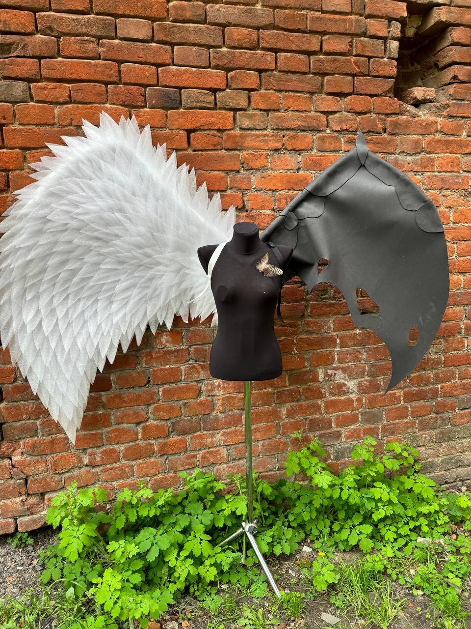 May include: A black mannequin wearing a pair of white angel wings and a pair of black devil wings. The mannequin is standing on a tripod stand in front of a brick wall.