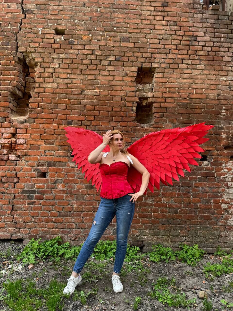 May include: A woman wearing a red corset and blue jeans with large red paper wings attached to her back. She is standing in front of a brick wall.