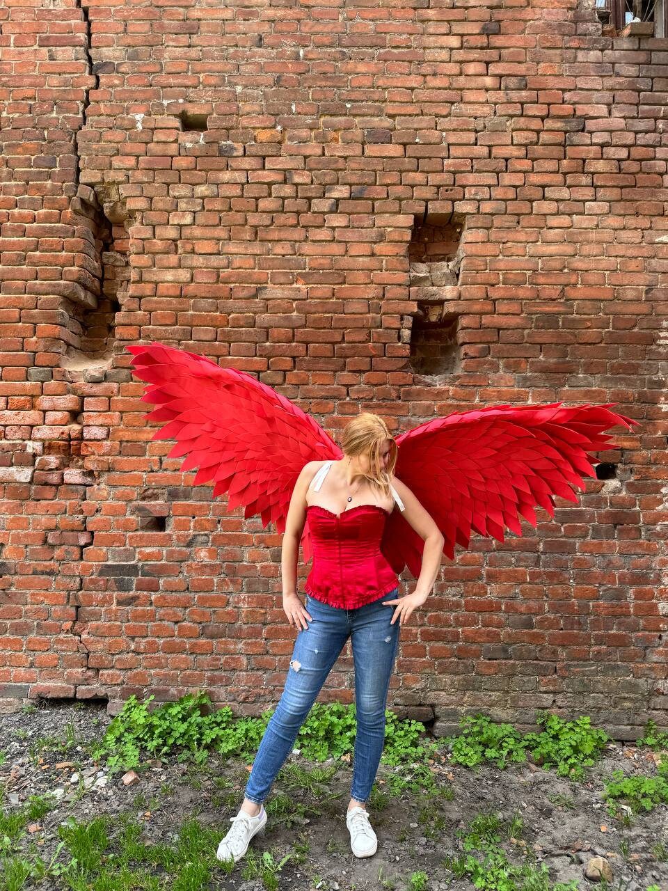 May include: A person wearing a red corset and blue jeans with large red feathered wings against a brick wall background.