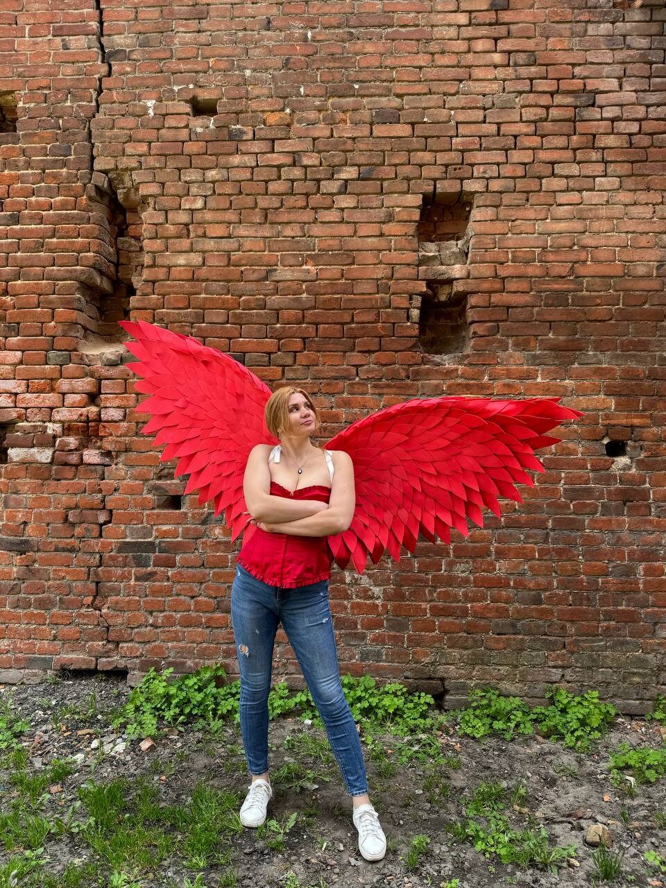 May include: A woman wearing a red top and blue jeans stands in front of a brick wall with large red wings behind her.
