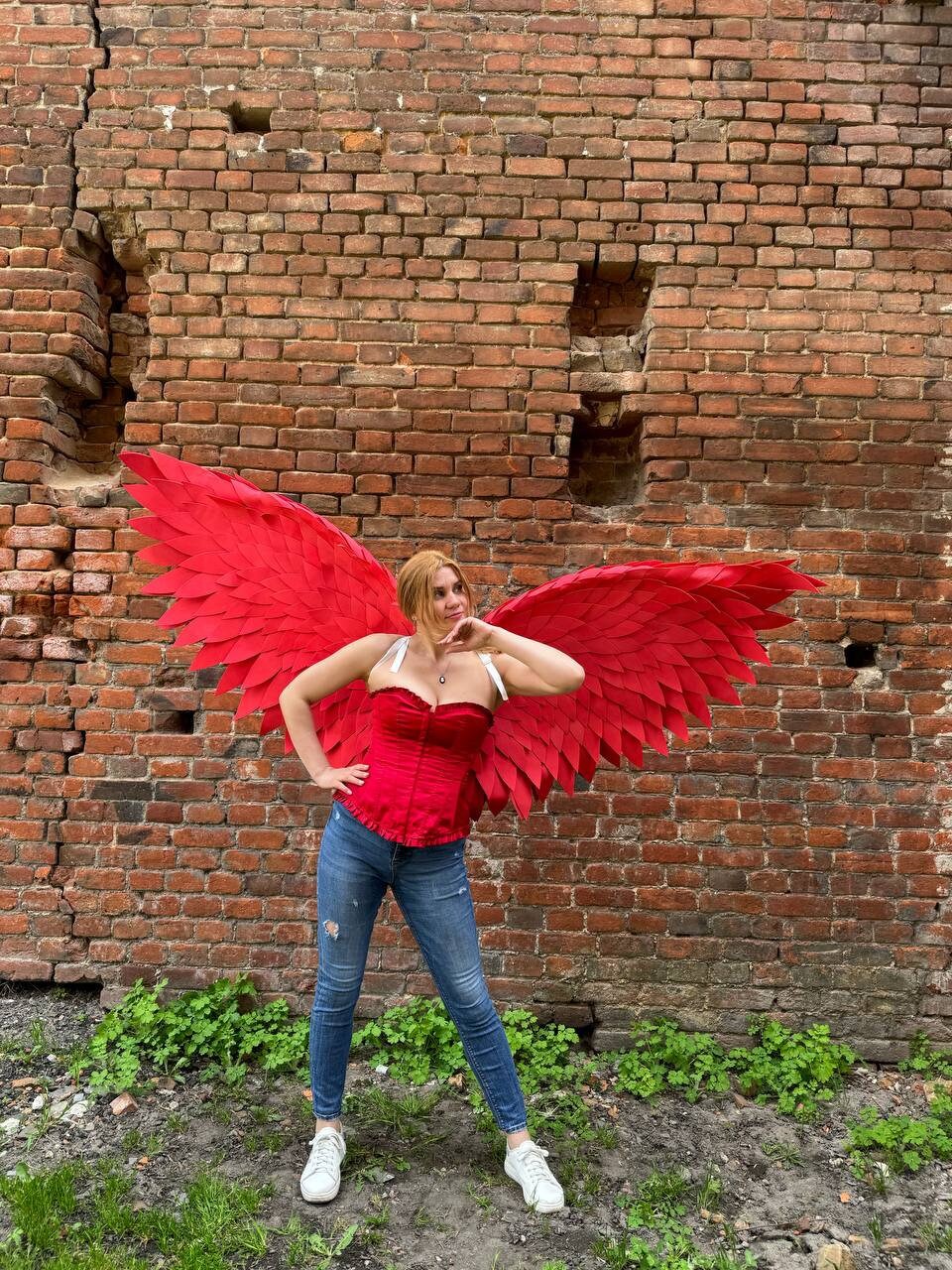May include: A woman wearing a red corset and blue jeans poses in front of a brick wall with large red feathered wings.