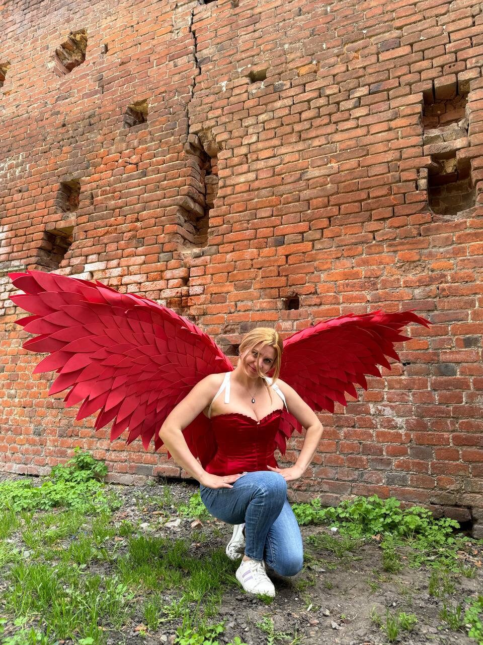 May include: A woman wearing a red corset and blue jeans poses with large red feathered wings in front of a brick wall.