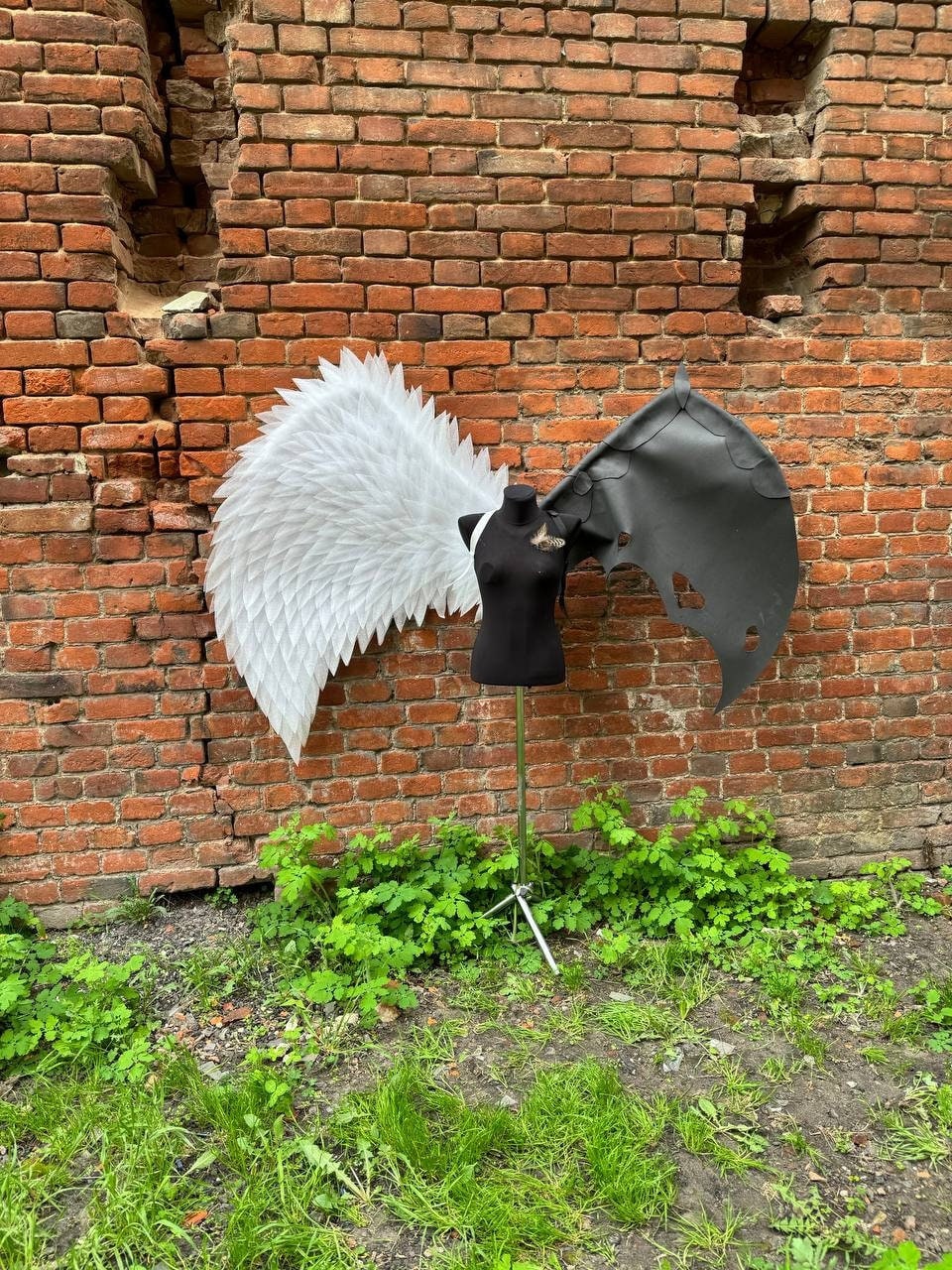 May include: A white angel wing and a black devil wing are displayed on a mannequin against a brick wall. The wings are made of feathers and fabric.