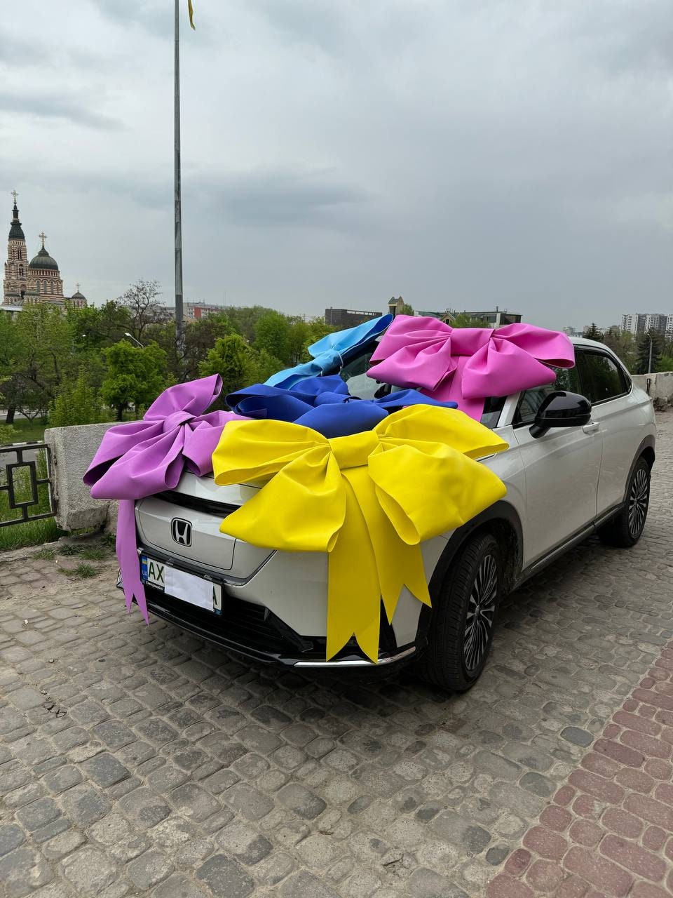 May include: A white car with a large yellow bow, a pink bow, and a blue bow on top. The car is parked on a cobblestone street.