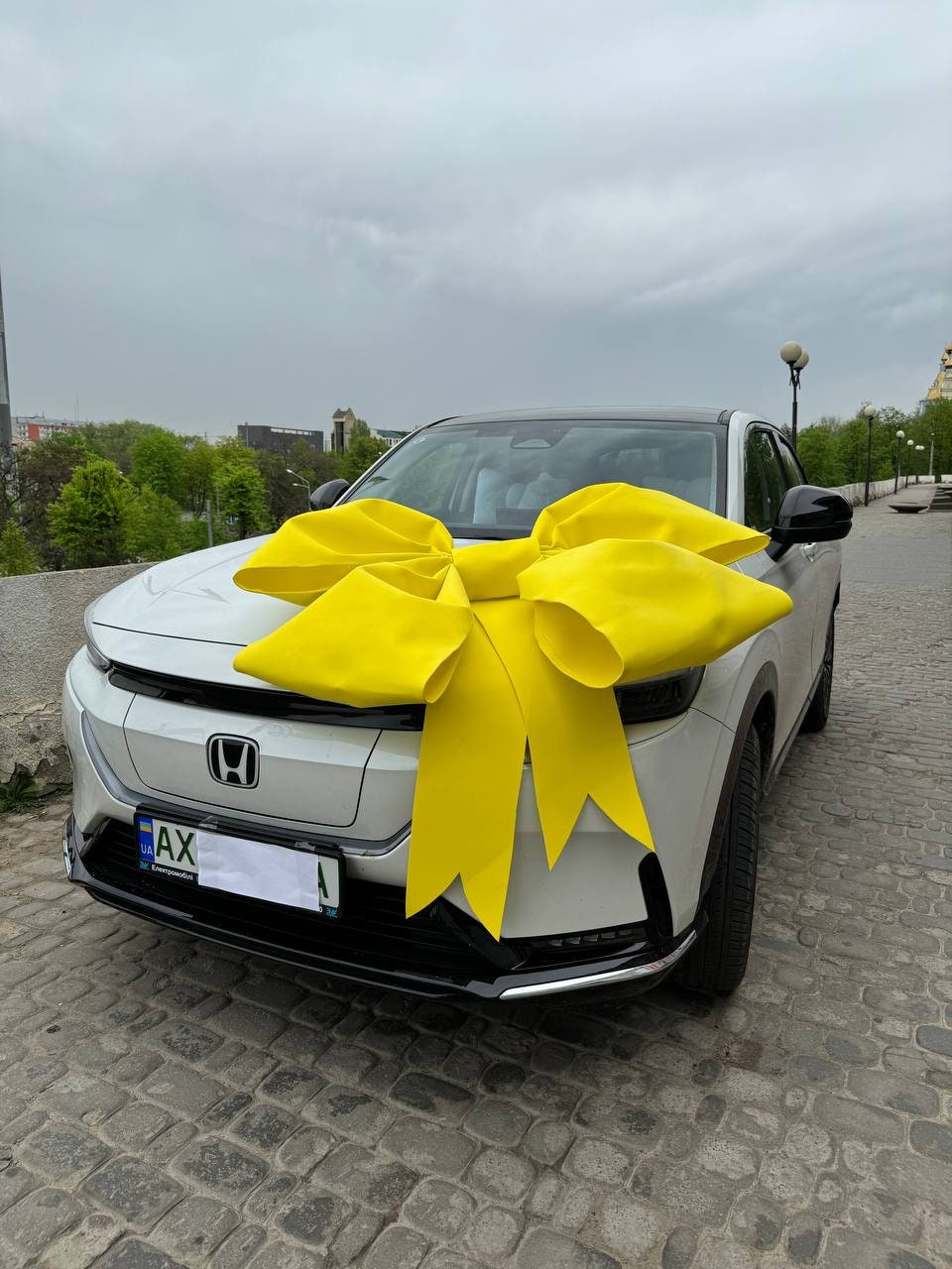 May include: A white car with a large yellow bow on the hood. The car has a license plate that reads "AX".