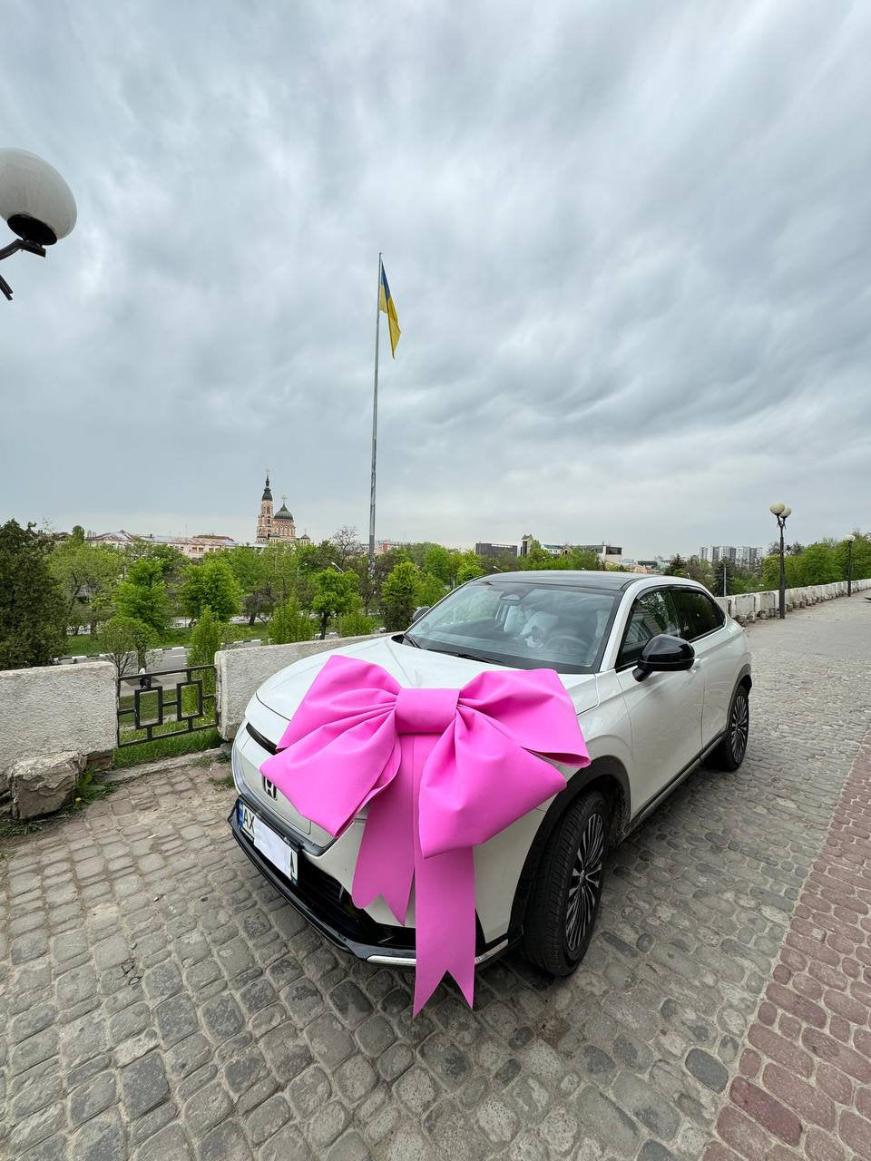 May include: A white car with a large pink bow on the hood. The car is parked on a cobblestone street with a Ukrainian flag flying in the background.