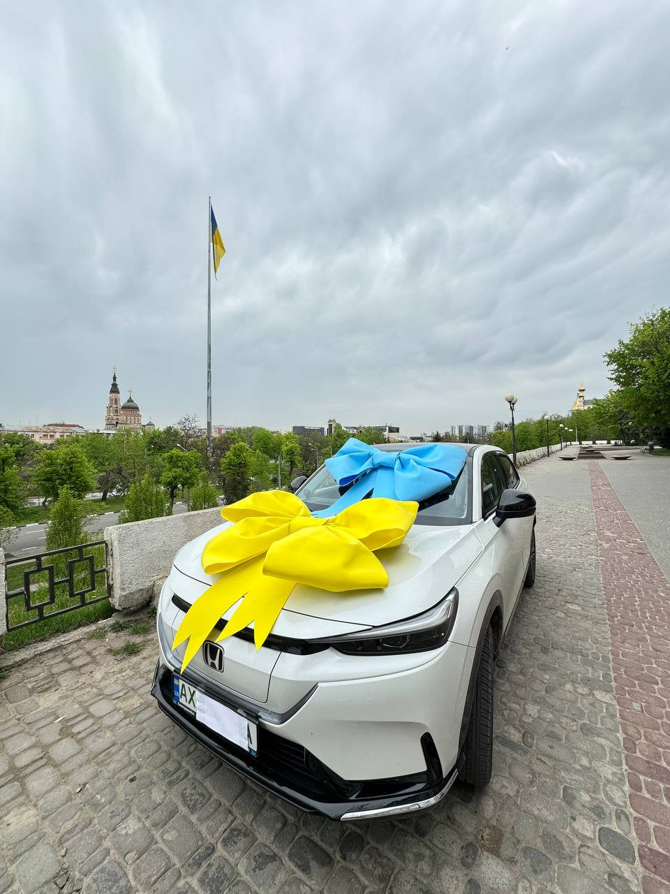 May include: A white car with a large yellow bow and a blue bow on the hood. The Ukrainian flag flies in the background. The car is parked on a brick-paved area.