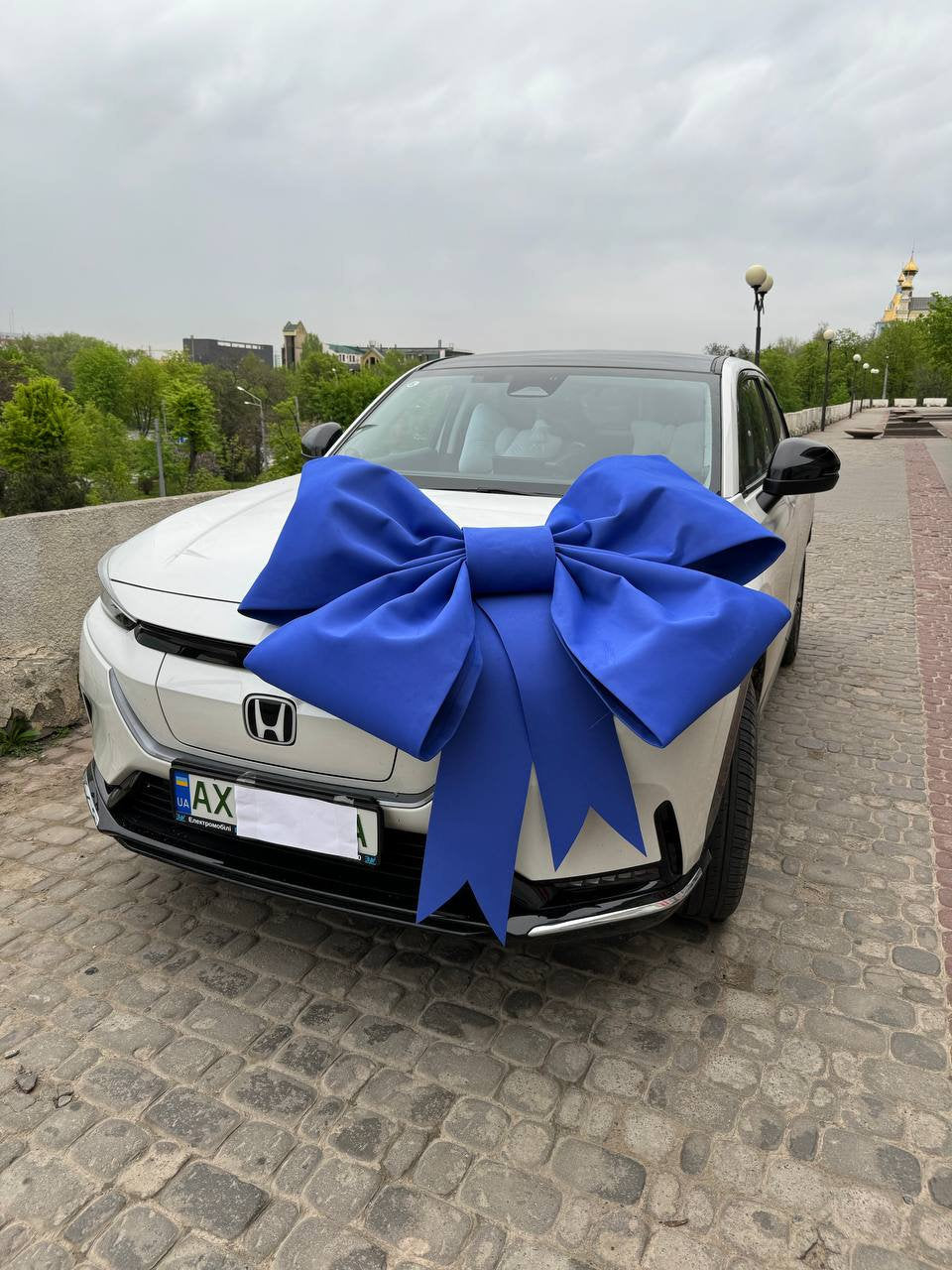 May include: A white car with a large blue bow on the hood. The car has a license plate that says "AX".