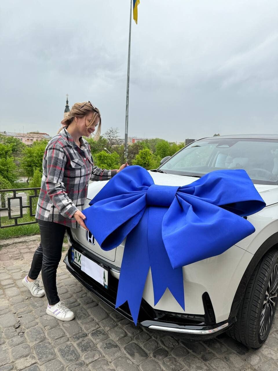 May include: A white car with a large blue bow on the hood. A woman in a plaid shirt and black pants stands next to the car.