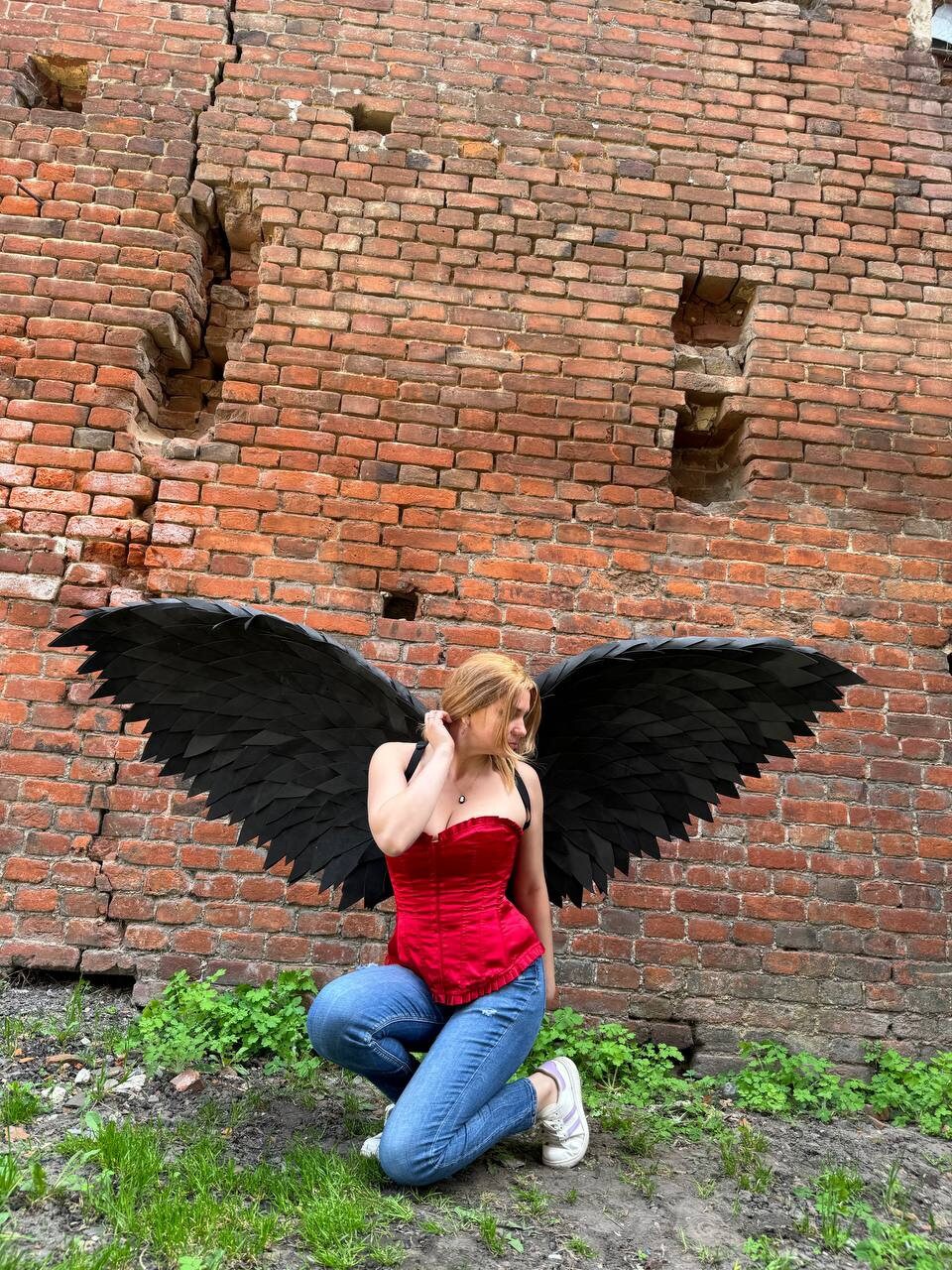 May include: A person wearing a red corset and blue jeans poses with large black feathered wings in front of a brick wall.