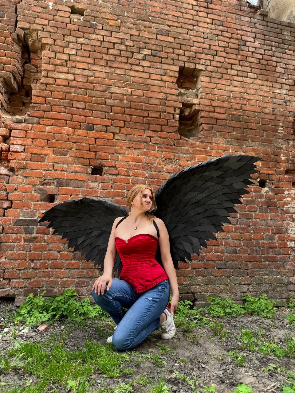 May include: A woman wearing a red corset and blue jeans poses with large black angel wings in front of a brick wall.