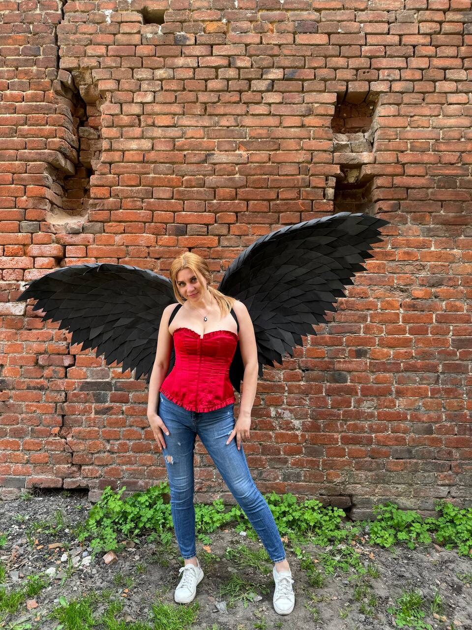 May include: A woman wearing a red corset and blue jeans with black angel wings against a brick wall background.