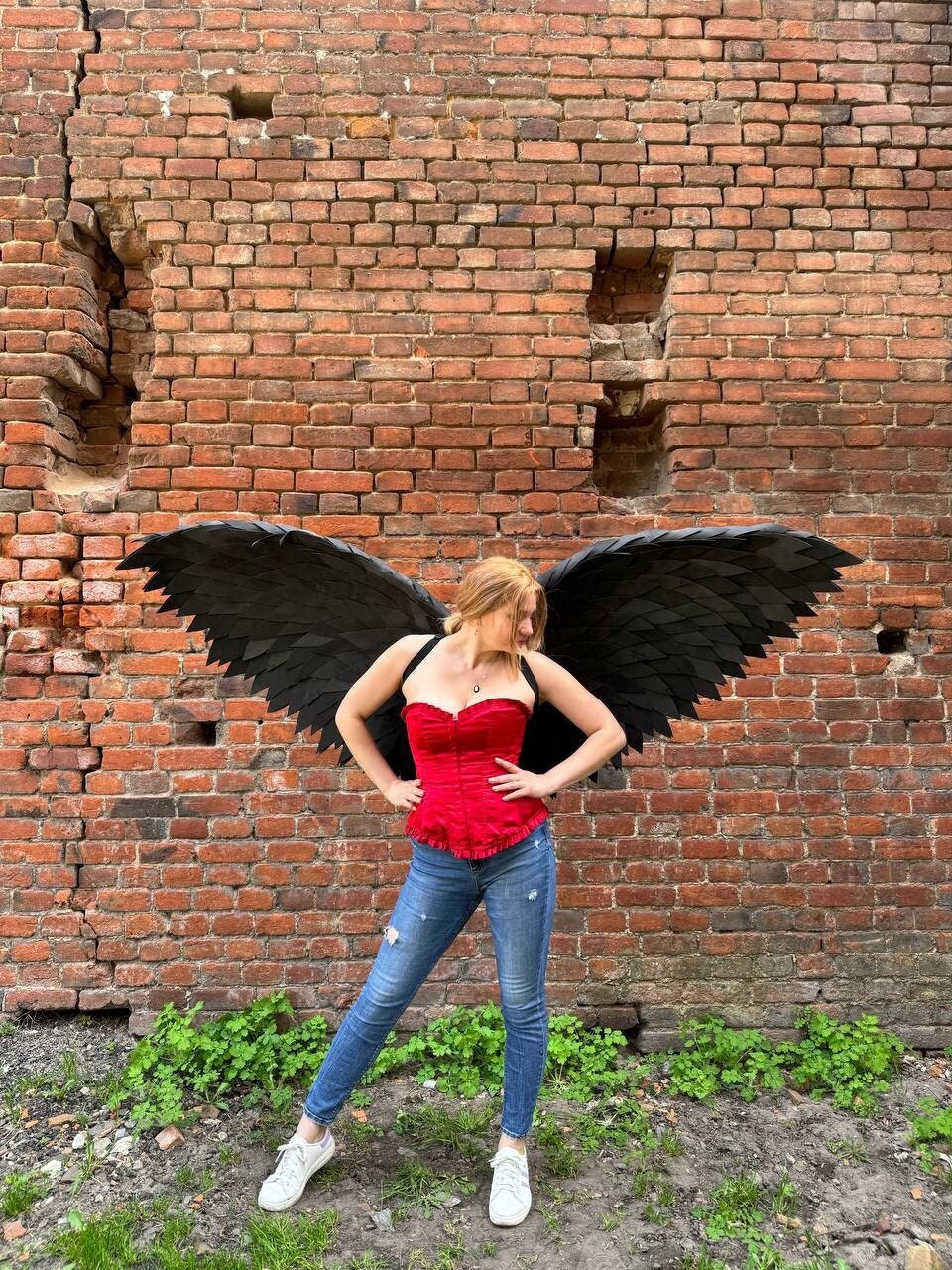 May include: A person wearing a red corset and blue jeans with a pair of large black angel wings.