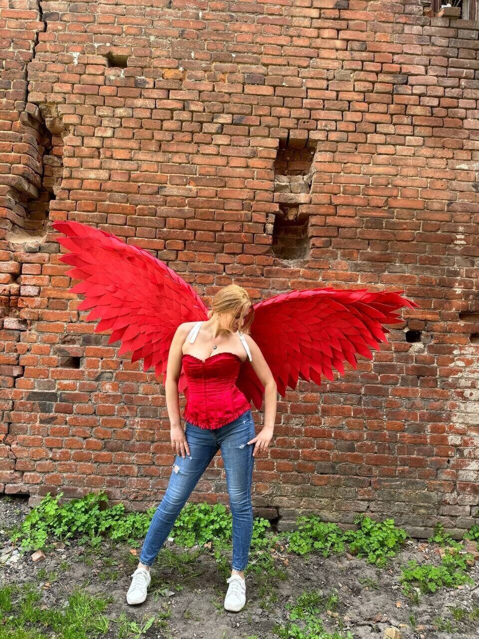 May include: A person wearing a red corset and blue jeans with large red feathered wings against a brick wall background.