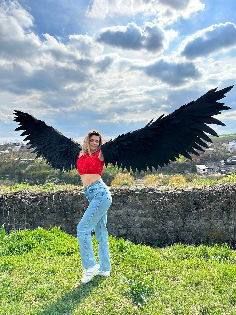 May include: A person wearing a red crop top and blue jeans is posing with large black feathered wings. The wings are spread wide and the person is looking at the camera. The background is a blue sky with white clouds.