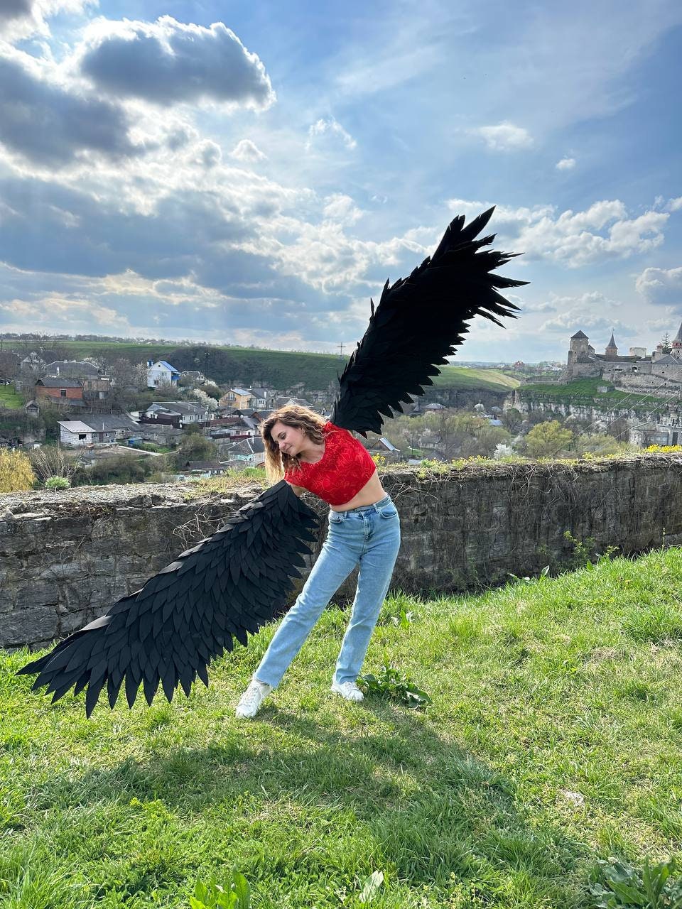May include: A person wearing large, black feathered wings, a red top, and blue jeans stands on a grassy area. The wings are spread wide against a backdrop of a cloudy sky and a distant town.