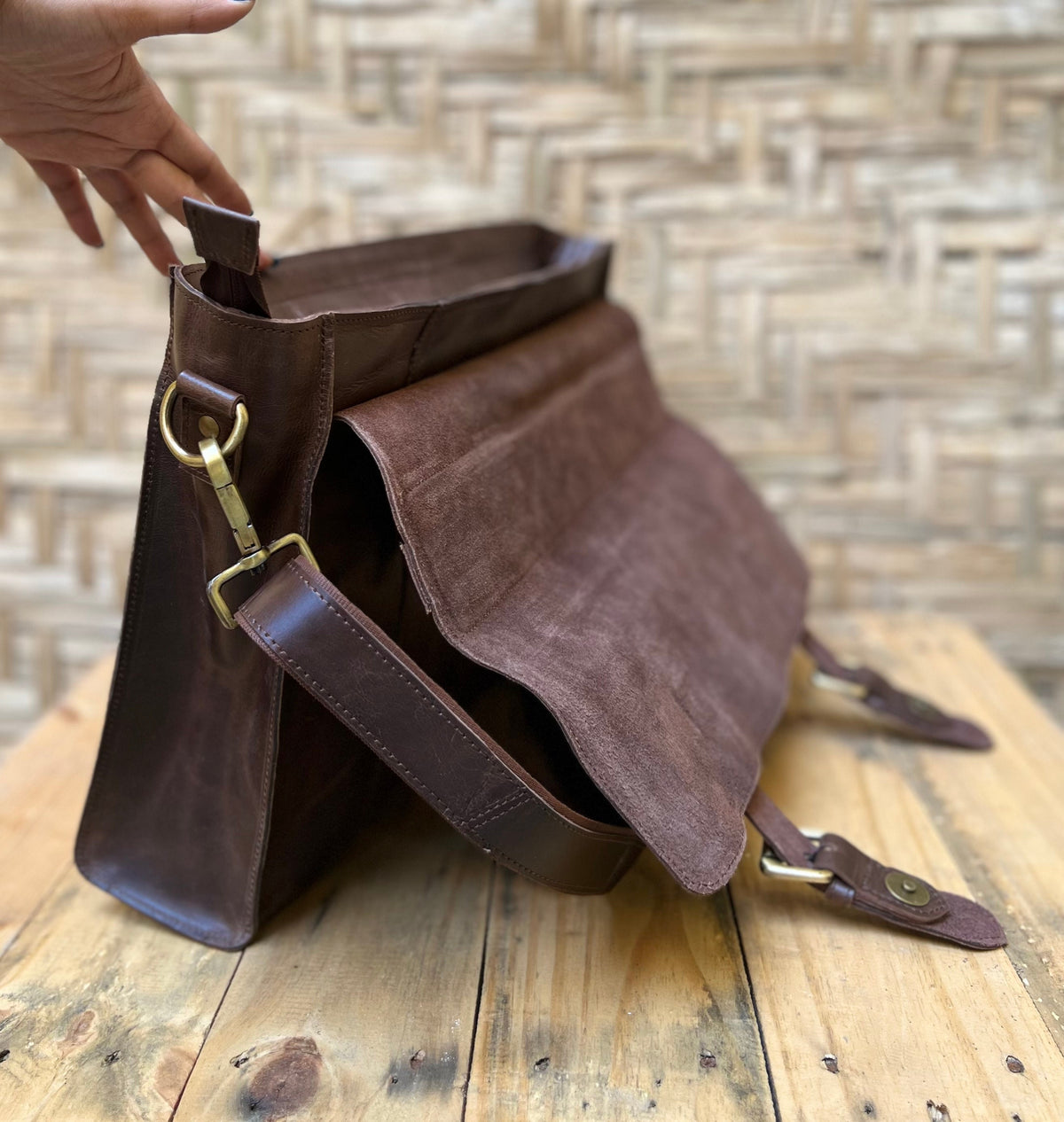 May include: A brown leather messenger bag with a suede flap. The bag features a shoulder strap, brass hardware, and a magnetic closure. The bag is open, revealing the interior. The bag is sitting on a wooden surface.