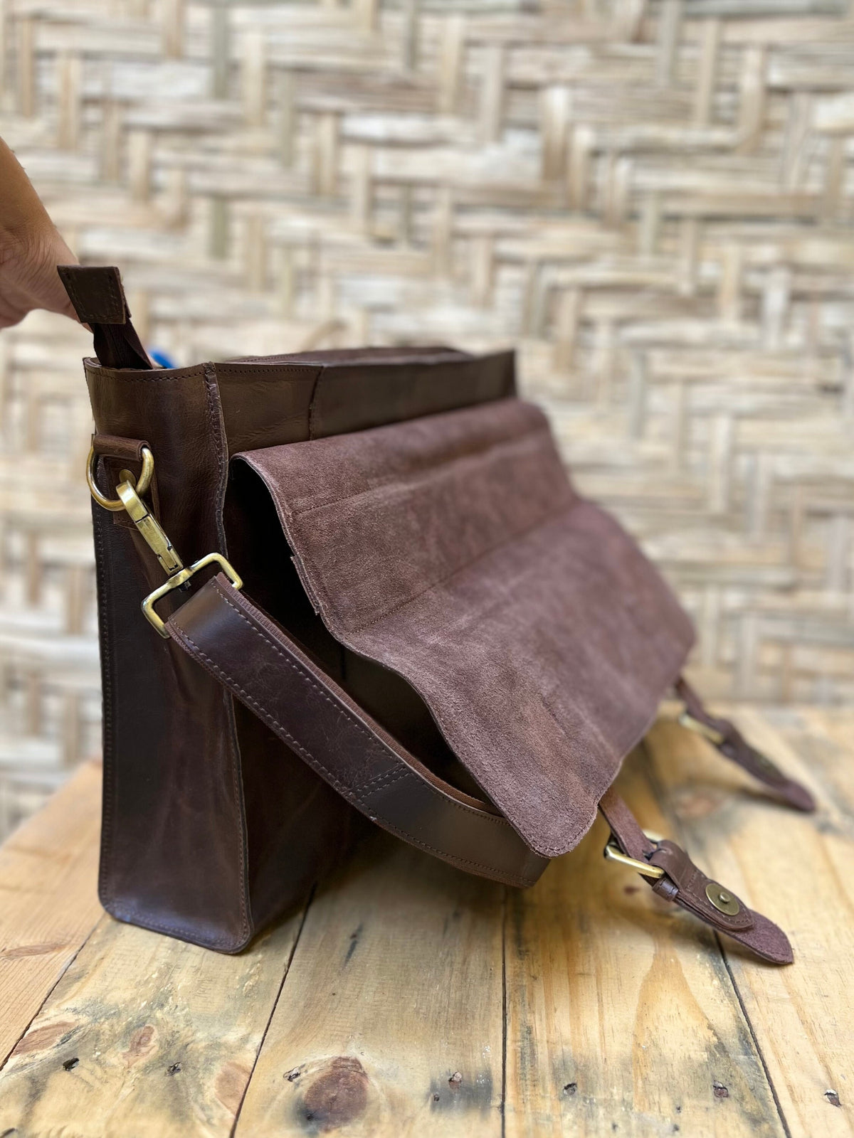 May include: A brown leather messenger bag with a suede flap and adjustable shoulder strap. The bag features brass-colored hardware and a rectangular shape. The bag is sitting on a wooden surface.