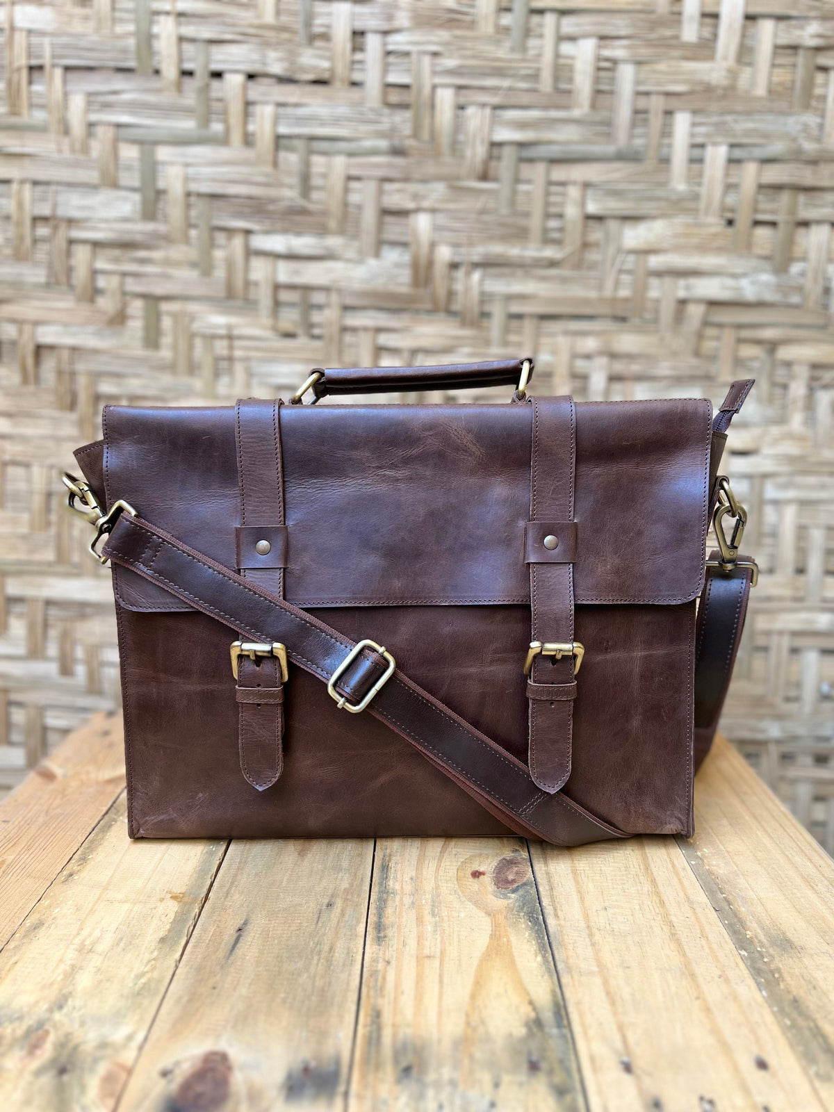 May include: A brown leather briefcase with a top handle, shoulder strap, and brass-colored hardware. The bag has two front straps with buckles and a flap closure. The leather has a distressed appearance, and the bag is sitting on a wooden surface.