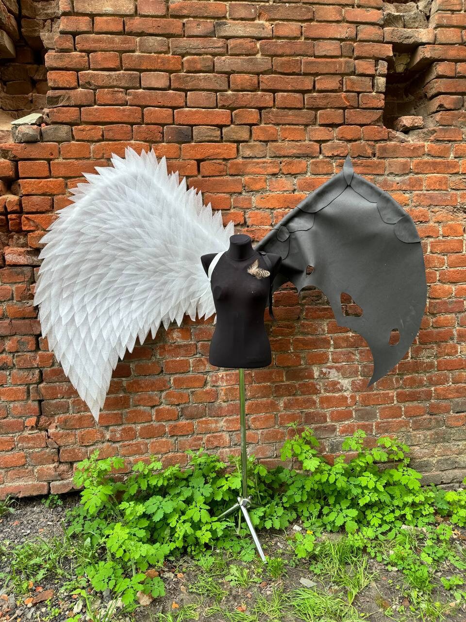 May include: A white angel wing and a black devil wing attached to a black mannequin. The wings are made of feathers and leather. The mannequin is standing in front of a brick wall.