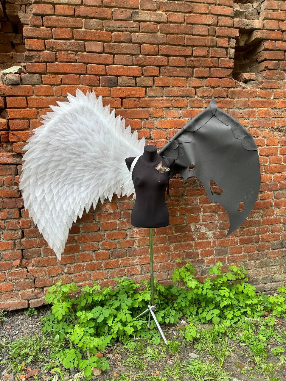 May include: A white feathered angel wing and a black leather demon wing are attached to a black mannequin. The mannequin is standing on a tripod stand in front of a brick wall.
