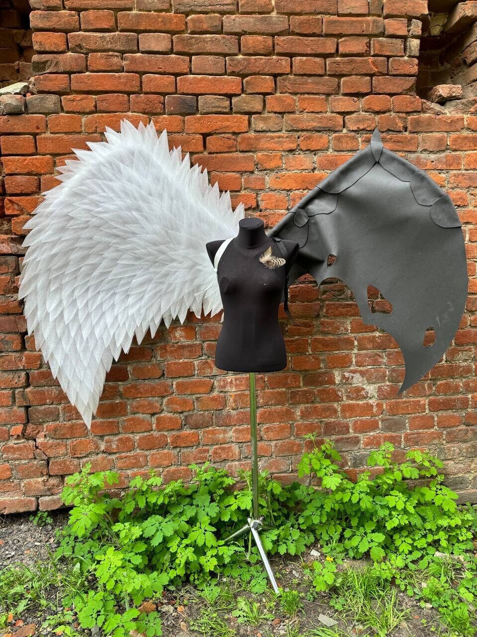 May include: A white feathered angel wing and a black leather bat wing are displayed on a mannequin against a brick wall. The wings are attached to the mannequin's back.