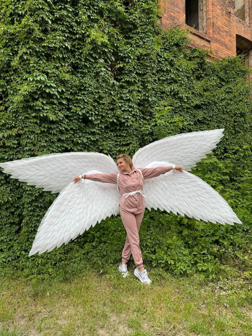 May include: A person wearing a pink jumpsuit and white angel wings, standing in front of a green ivy covered wall.