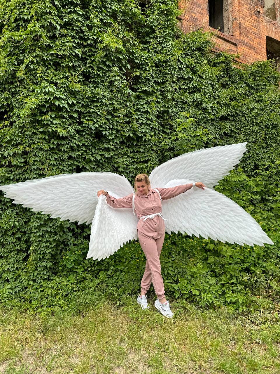 May include: A person wearing a pink jumpsuit and white sneakers is holding large white angel wings in front of a green ivy wall.