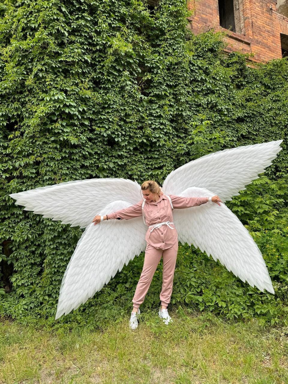 May include: A person wearing a pink jumpsuit and white sneakers is standing in front of a green wall with large white angel wings.