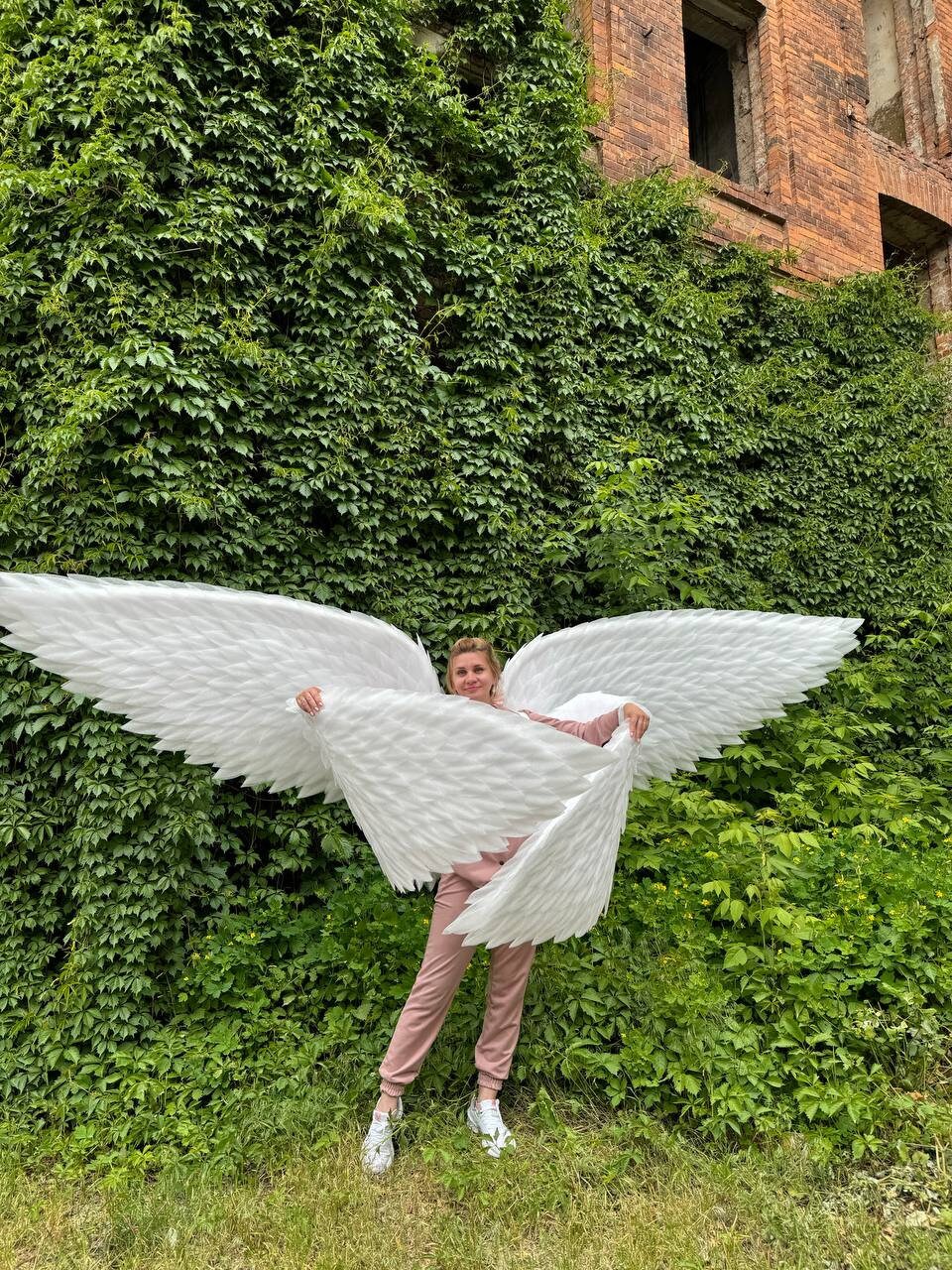 May include: A person wearing a pink jumpsuit and white sneakers is holding large white feathered angel wings in front of a green ivy covered wall.