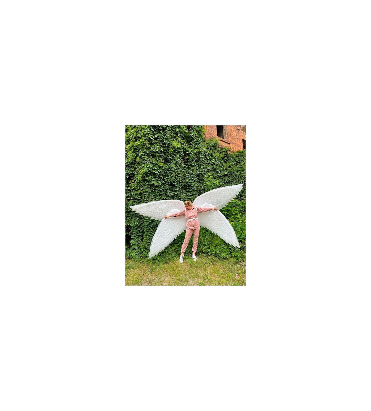 May include: A person wearing a pink outfit with white angel wings spread out behind them. The wings are large and feathery, and the person is standing in front of a green leafy wall.