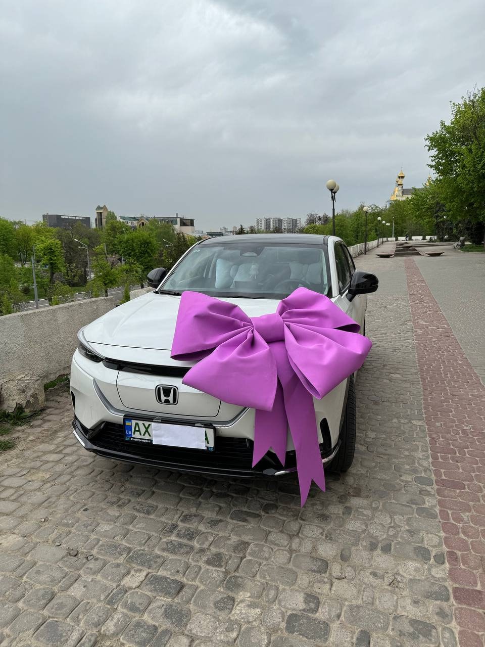 May include: A white car with a large, vibrant purple bow on the hood. The bow is the focal point, suggesting a celebratory or gift-giving occasion. The car is parked on a cobblestone street, with a cityscape in the background.