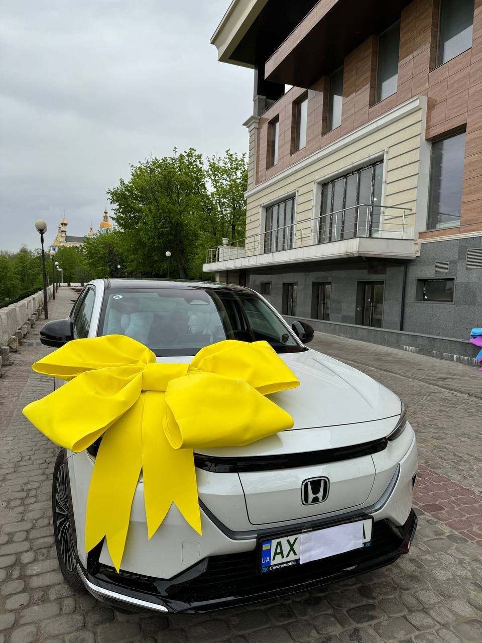 May include: A white car with a large yellow bow on the hood. The car has a license plate that says "UA AX".