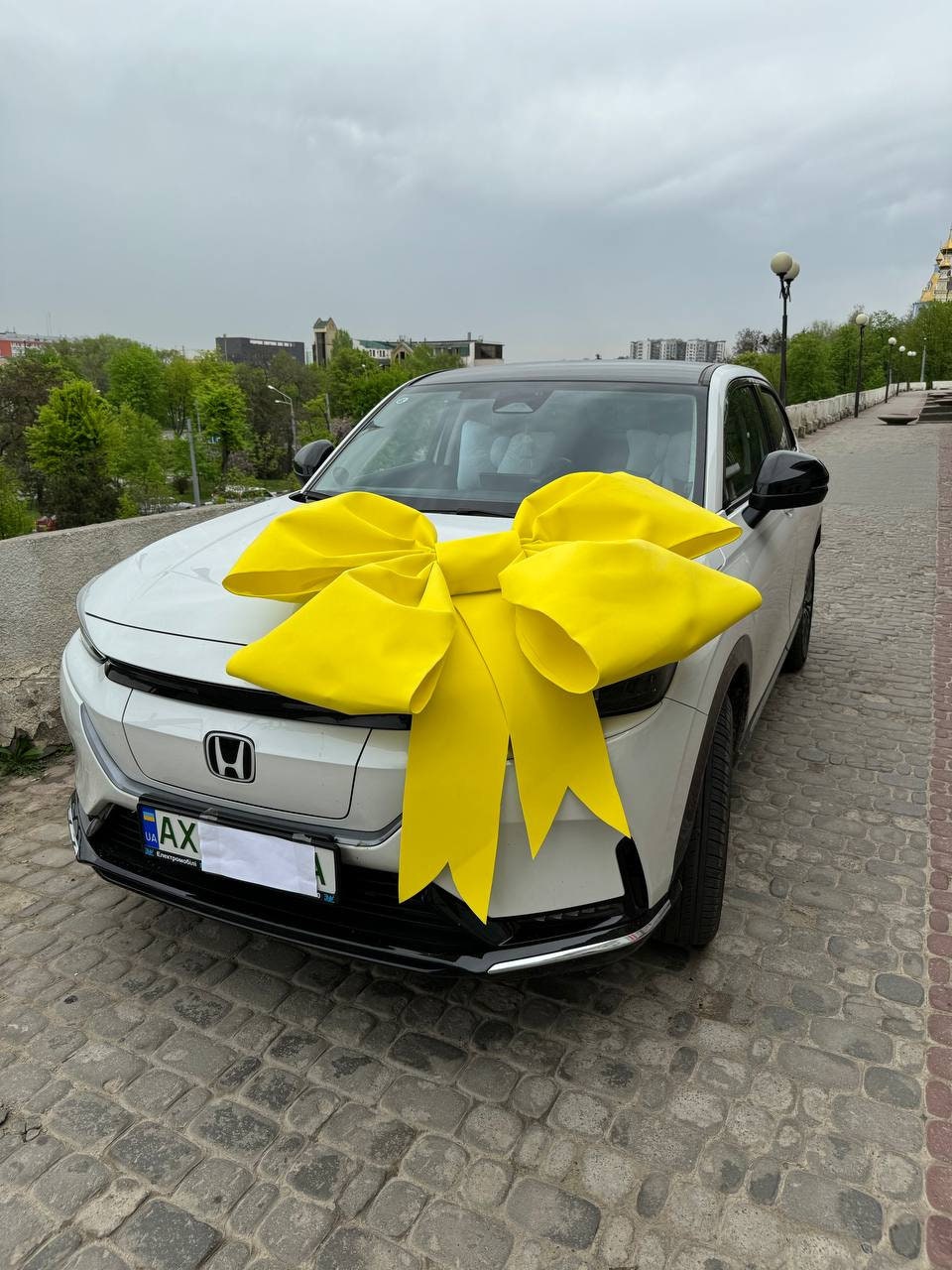 May include: A white car with a large yellow bow on the hood. The car has a license plate that says "AX".