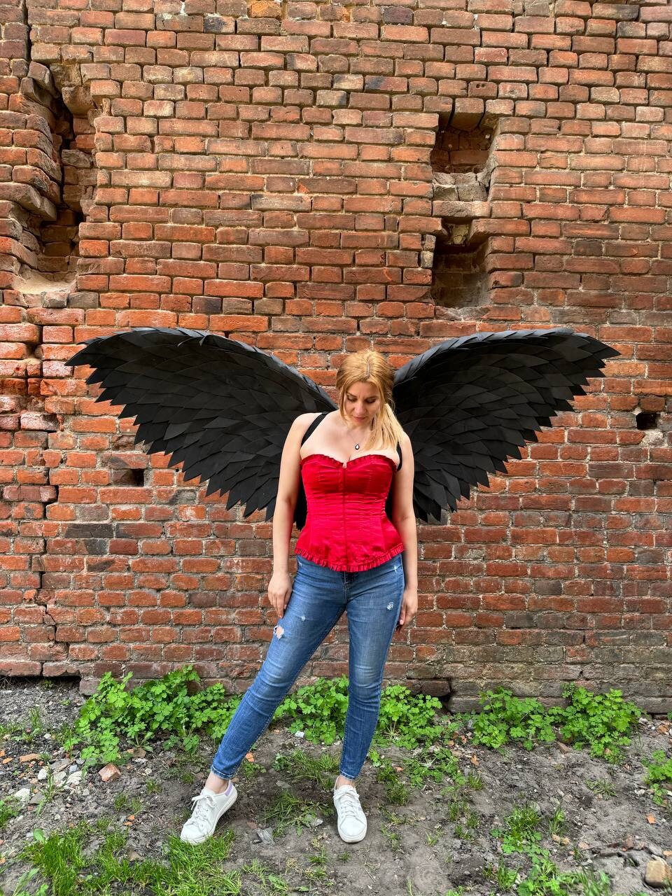 May include: A person wearing a red corset and blue jeans with black angel wings. The person is standing in front of a brick wall.