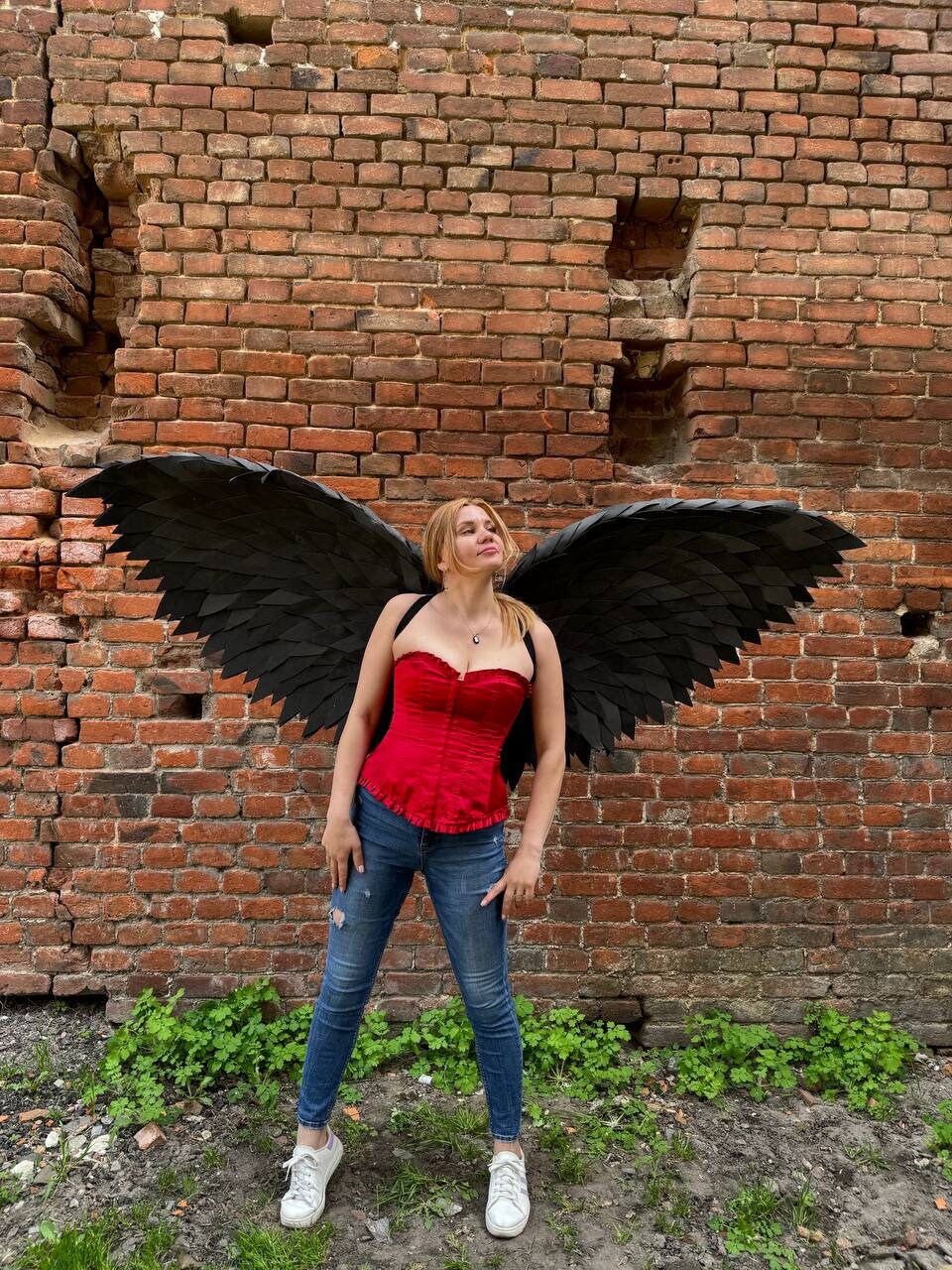 May include: A woman wearing a red corset and blue jeans stands in front of a brick wall with large black wings.