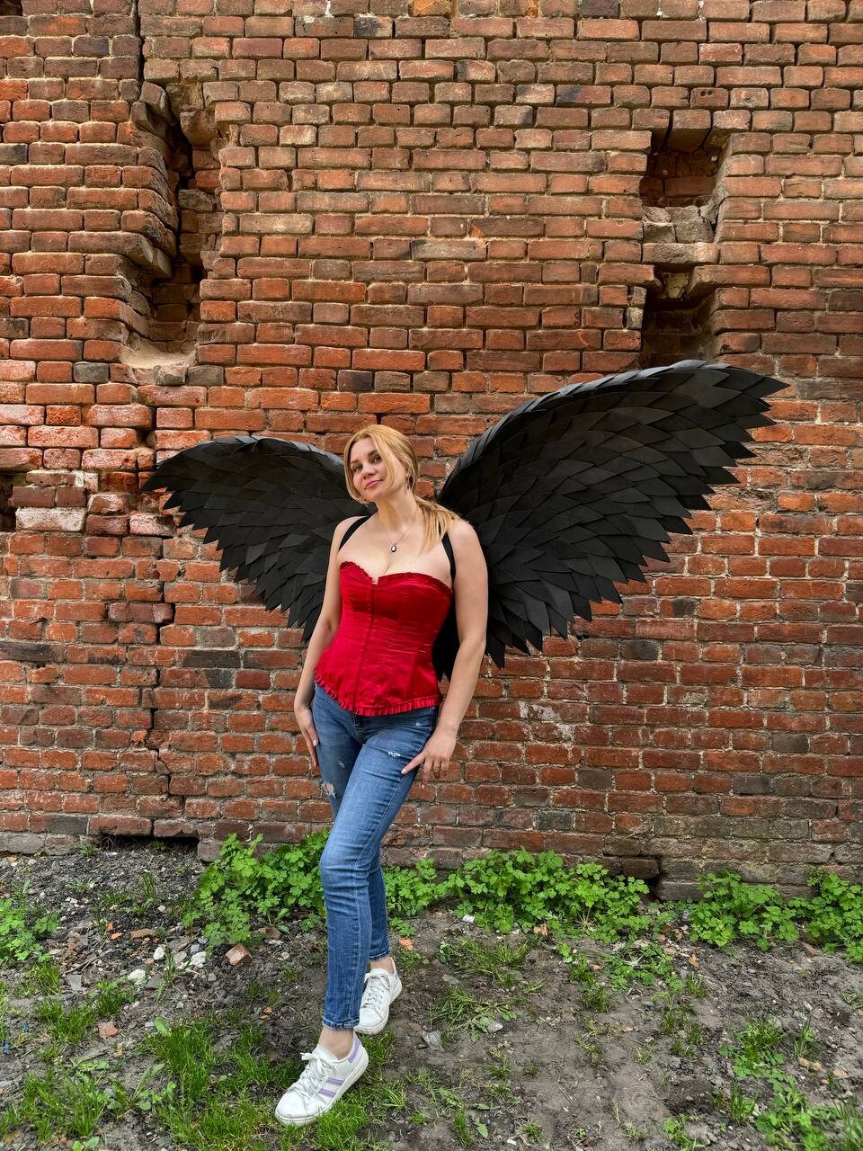 May include: A person wearing a red corset and blue jeans with large black feathered wings against a brick wall background.