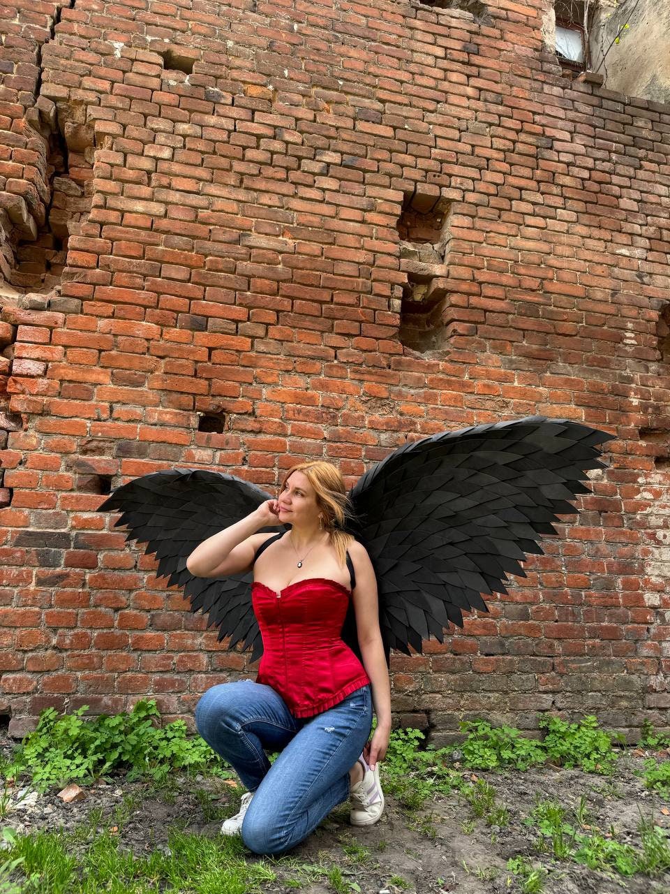 May include: A person wearing a red corset and blue jeans poses with large black wings in front of a brick wall.