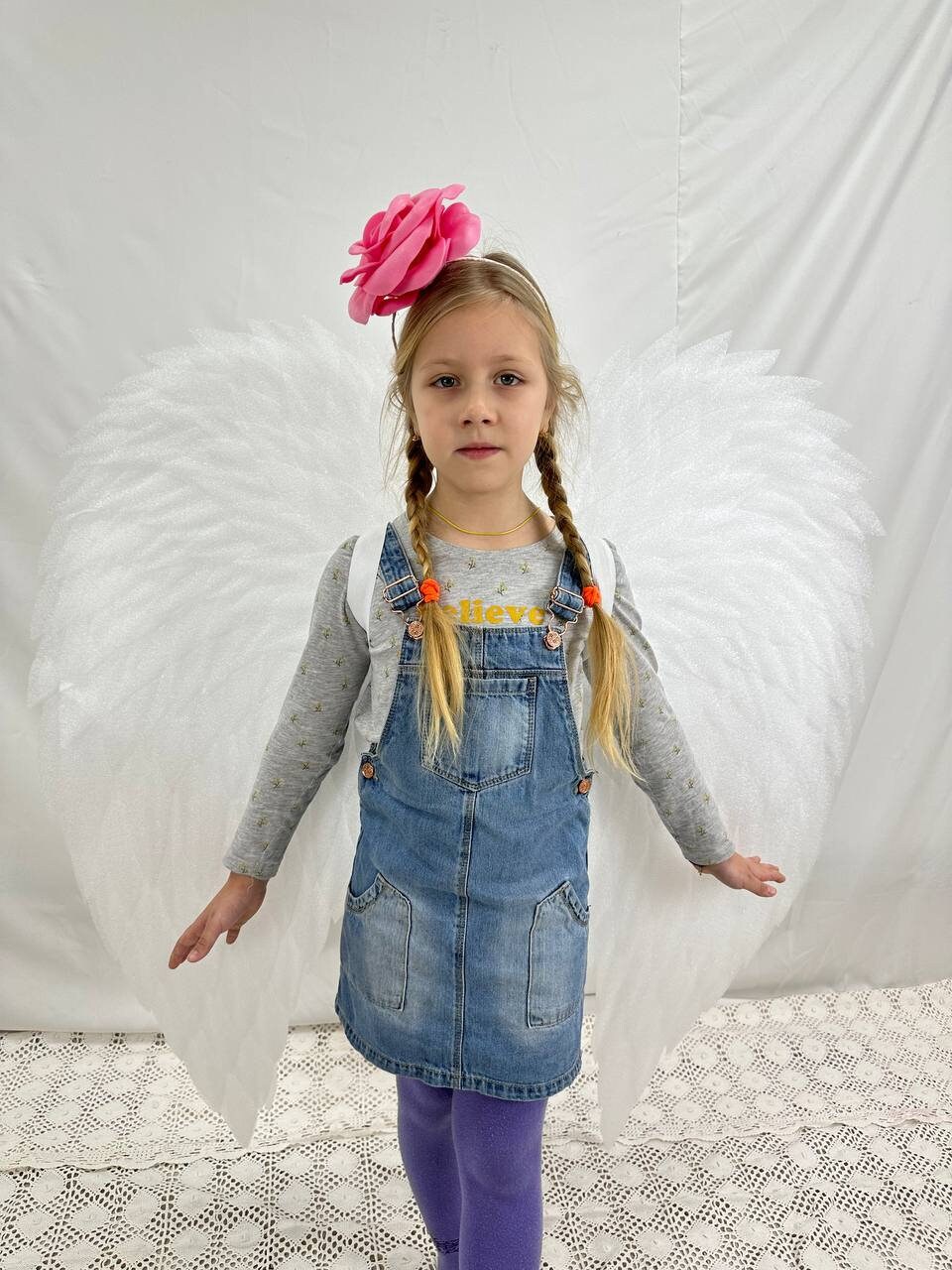 May include: A young girl wearing a denim overall dress and white angel wings with a pink flower headband. She has blonde hair in pigtails with orange hair ties. The background is a white wall with a white crocheted surface.
