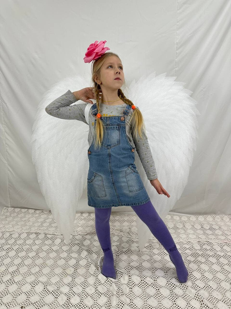 May include: A young girl wearing a denim overall dress and purple tights poses with large white angel wings. She has a pink flower headband in her hair and is looking up.
