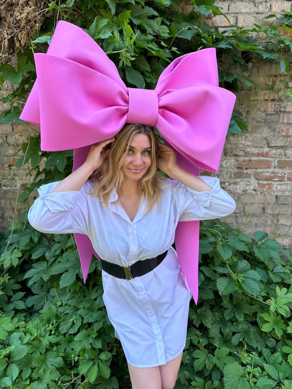 May include: A large, bright pink bow headpiece. The bow is worn by a person wearing a white button-down shirt and a black belt. The background features green foliage and a brick wall.