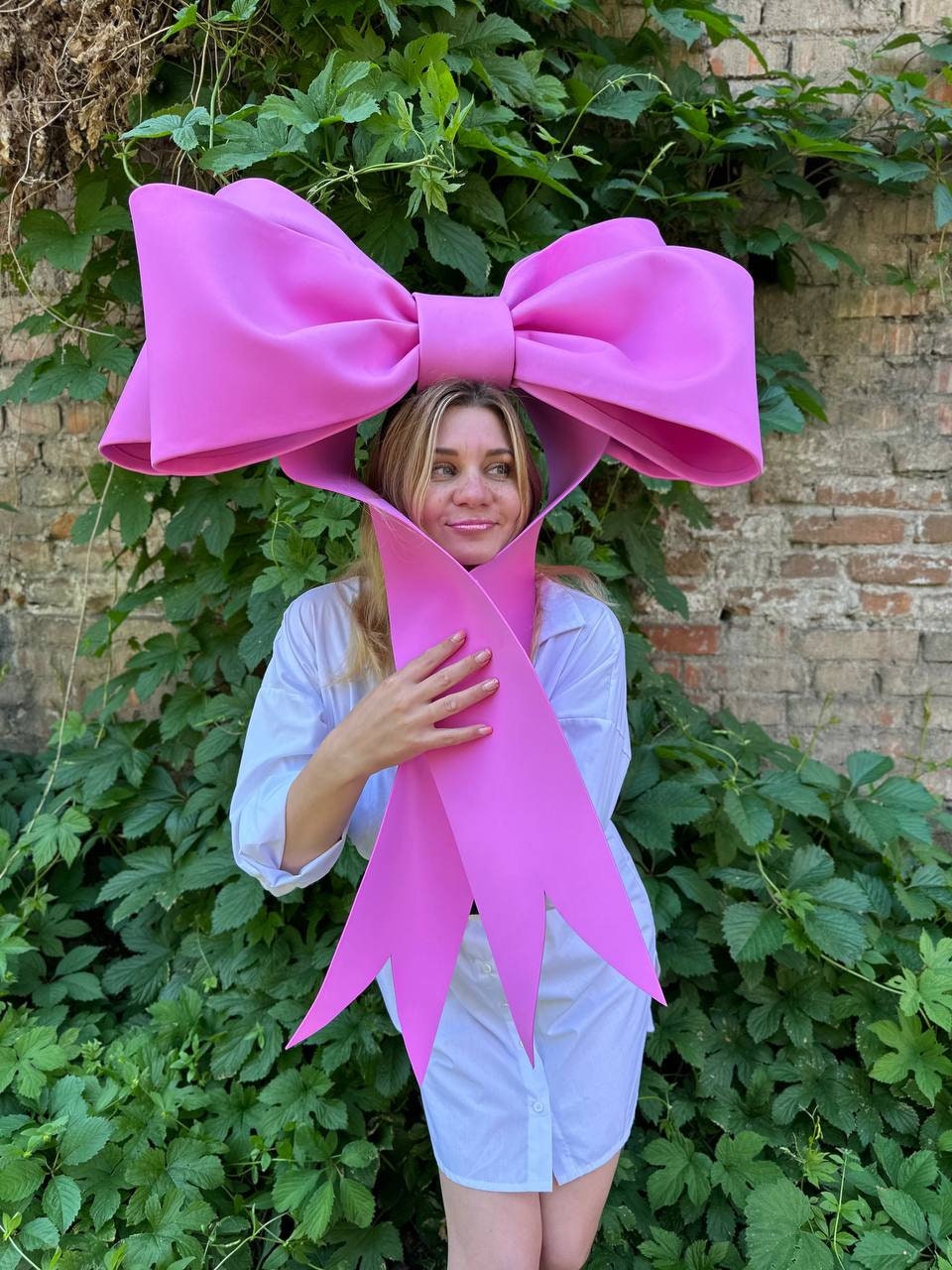 May include: A large, bright pink bow, worn as a headpiece. The bow is made of a thick material and has a classic bow shape. The person is wearing a white button-down shirt. The background is a brick wall and green foliage.