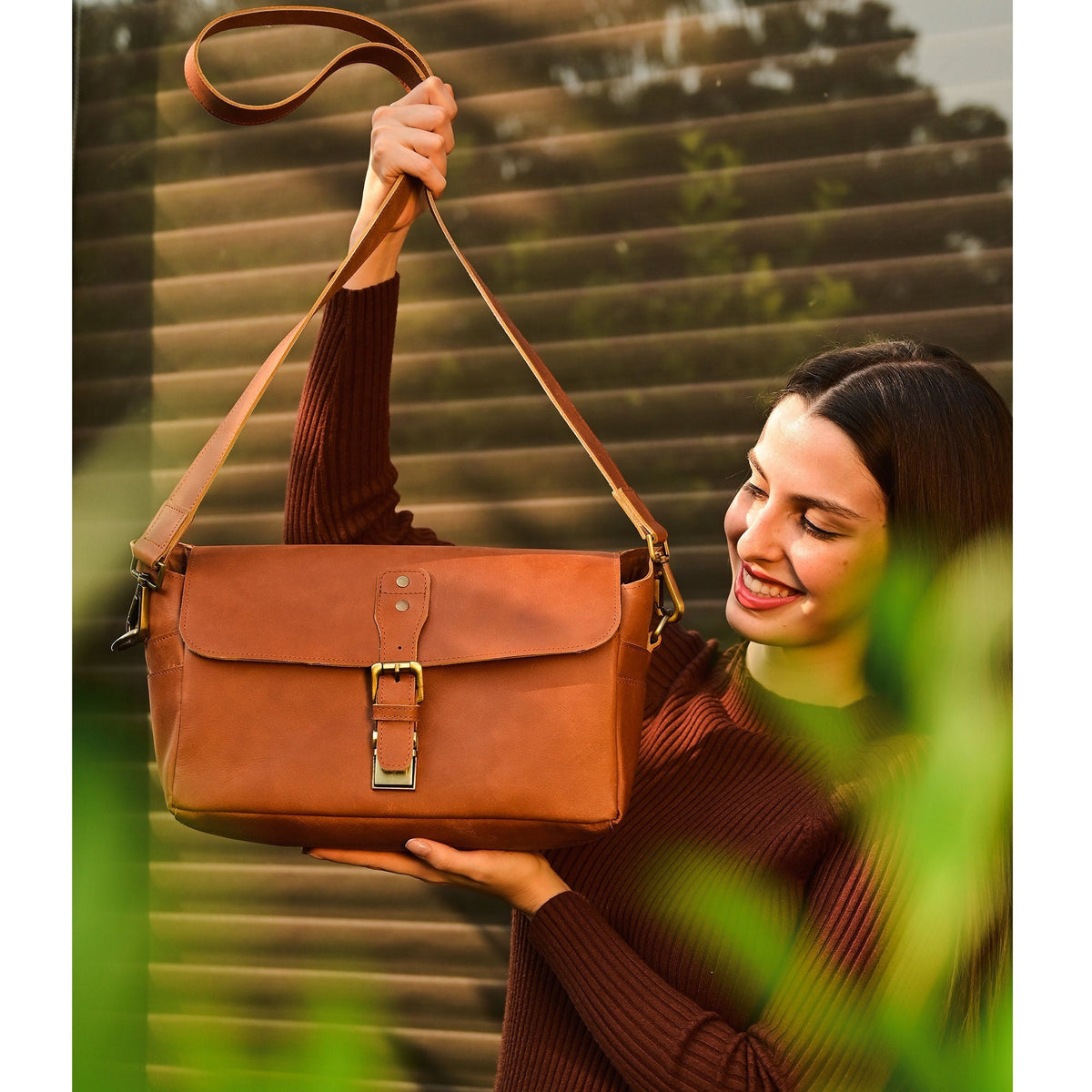 May include: A brown leather satchel bag with a brass buckle closure. The bag has a long adjustable strap and is being held by a person.
