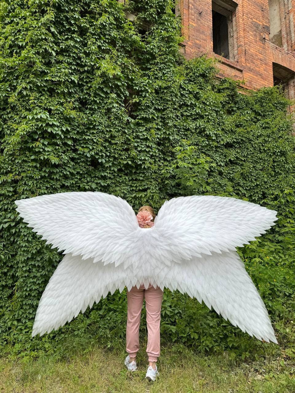 May include: A person wearing pink pants and white shoes is holding large white angel wings in front of a green ivy covered wall.