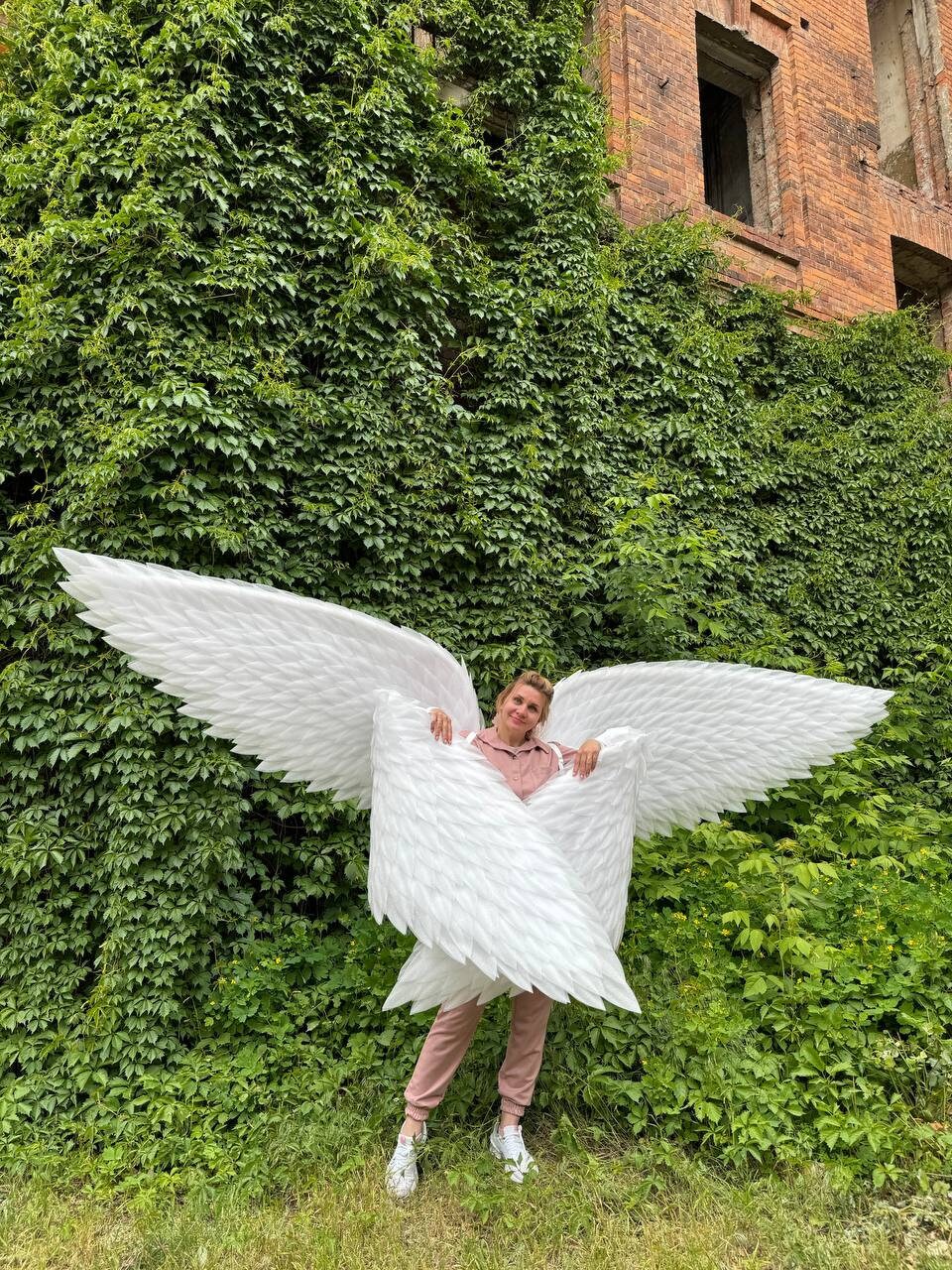 May include: A person wearing a pink shirt and white sneakers is holding large white angel wings in front of a green ivy covered wall.