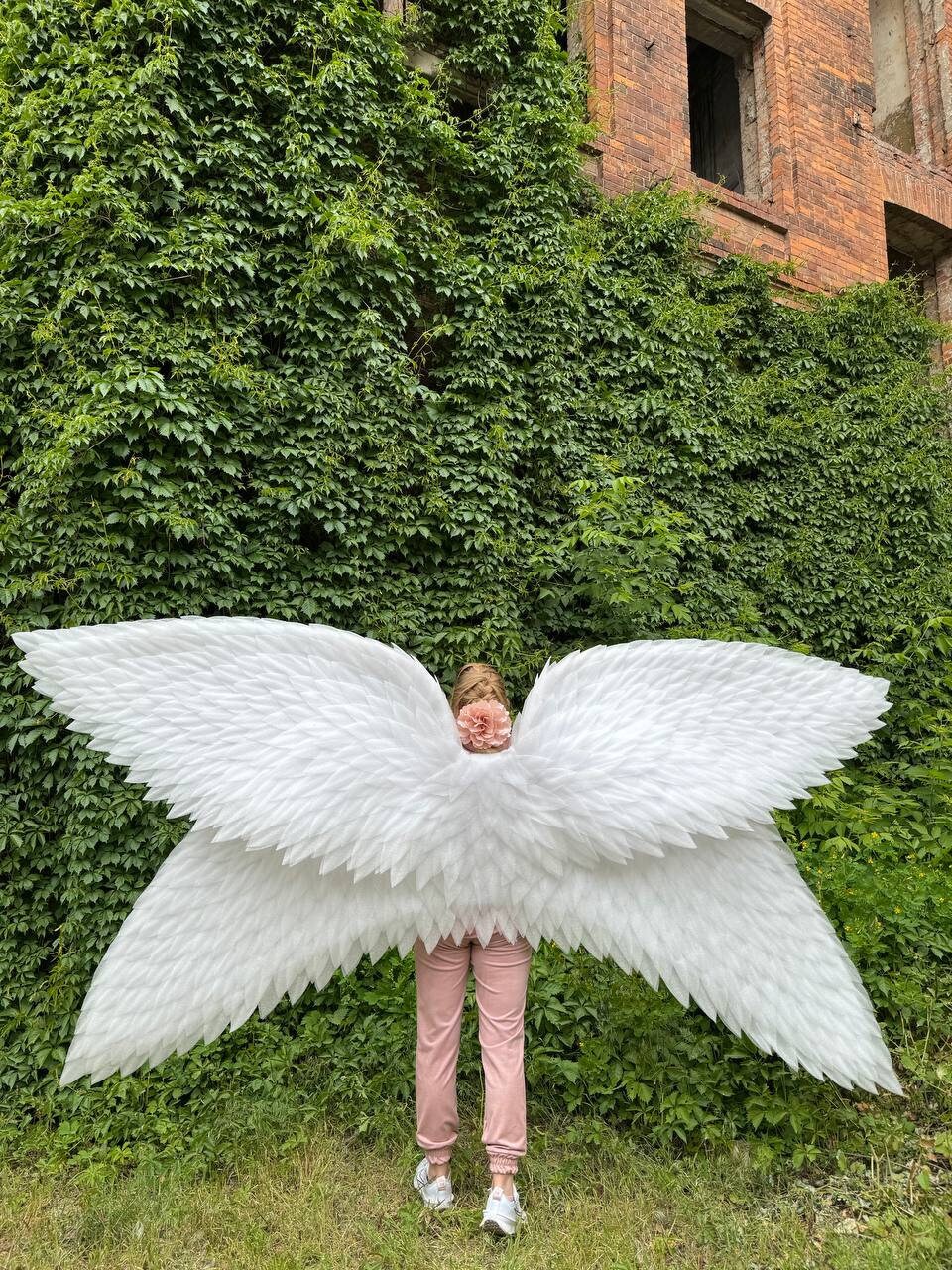 May include: A person wearing pink pants and white shoes is standing in front of a green ivy covered wall. They are wearing large white angel wings.